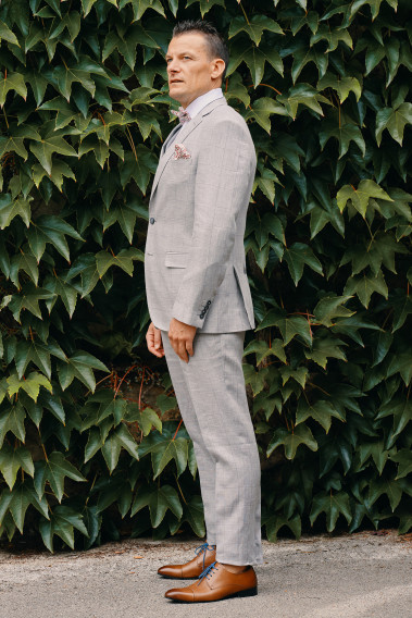 Homme de profil en costume SUAT gris à carreaux 3 pièces dans une cours. Il porte des chaussures marrons.