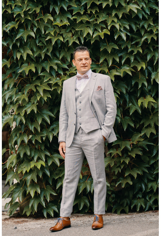 Homme de face en costume DIANA gris à carreaux 3 pièces dans une cours. Il porte des chaussures marrons.