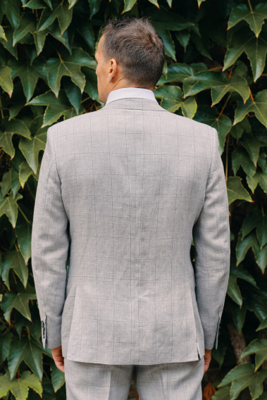 Homme de dos en costume DIANA gris à carreaux 3 pièces dans une cours. Il porte des chaussures marrons.
