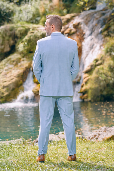 Homme de dos en costume SUAT couleur bleu pastel 3 pièces près d'une cascade. Il porte des chaussures marrons.