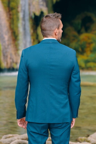 Homme de dos en costume SUAT couleur bleu-vert 3 pièces vers une cascade. Il porte des chaussures marrons.