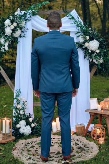 Homme de dos en costume SUAT couleur vert-bleu 3 pièces dans une cours . Il porte des chaussures marrons.