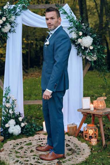 Homme de dos en costume SUAT couleur vert-bleu 3 pièces dans une cours. Il porte un nœud papillon a motif et un pins.