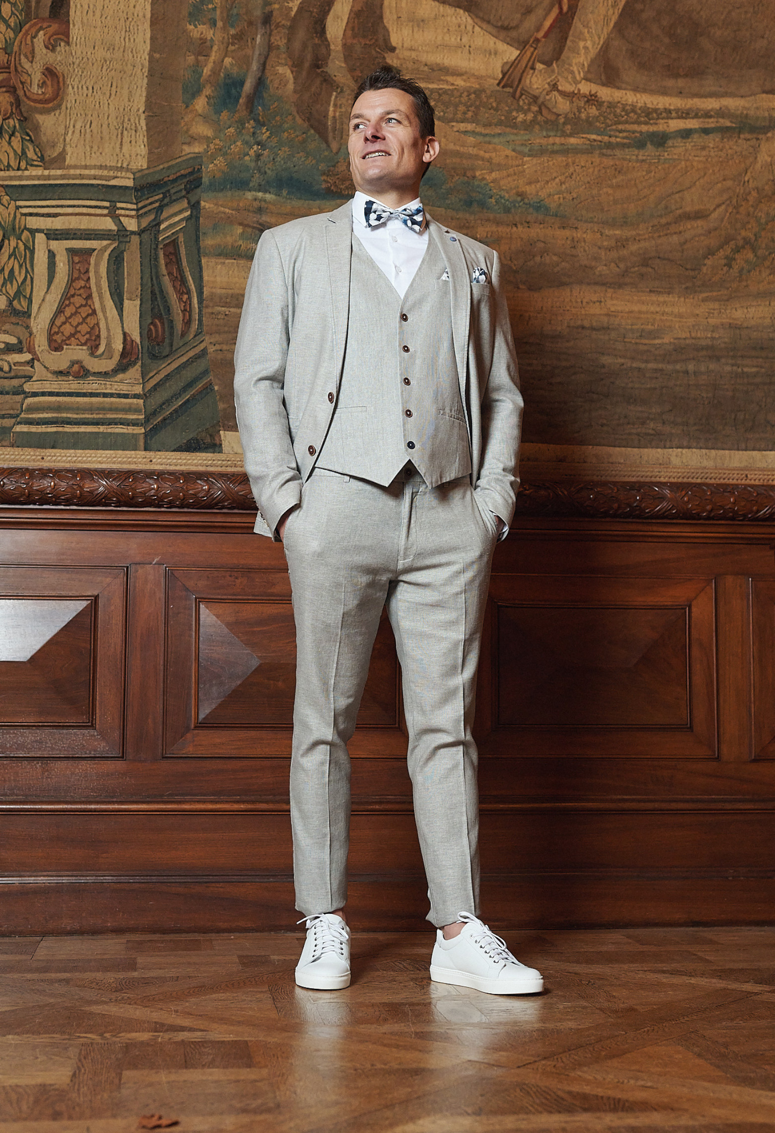 Homme de face en costume GIORGIOC couleur beige 3 pièces. Il porte un nœuds papillon noir à motif et des baskets blanches.