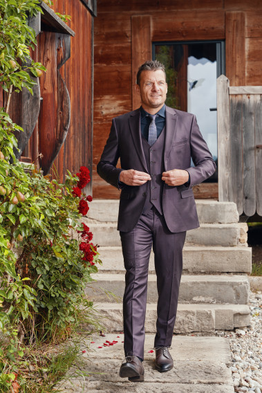 Homme de face en costume CITIZEN bordeaux 2 pièces devant un chalet.