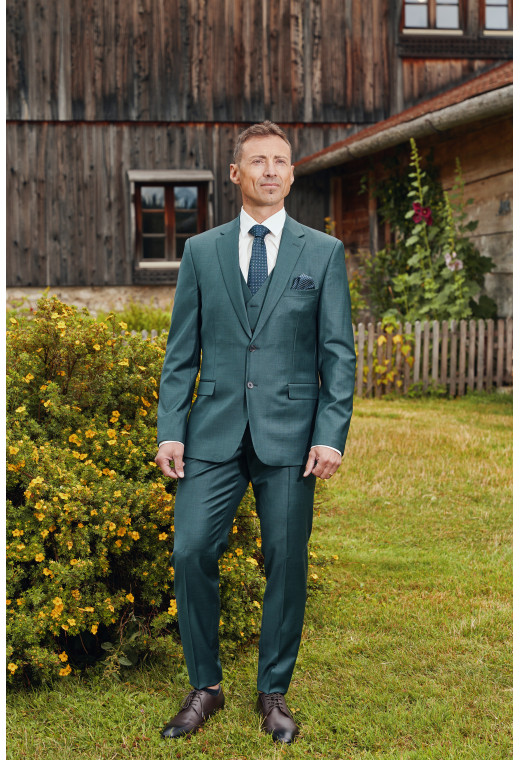 Homme de face en costume CITIZEN vert 2 pièces devant un chalet.