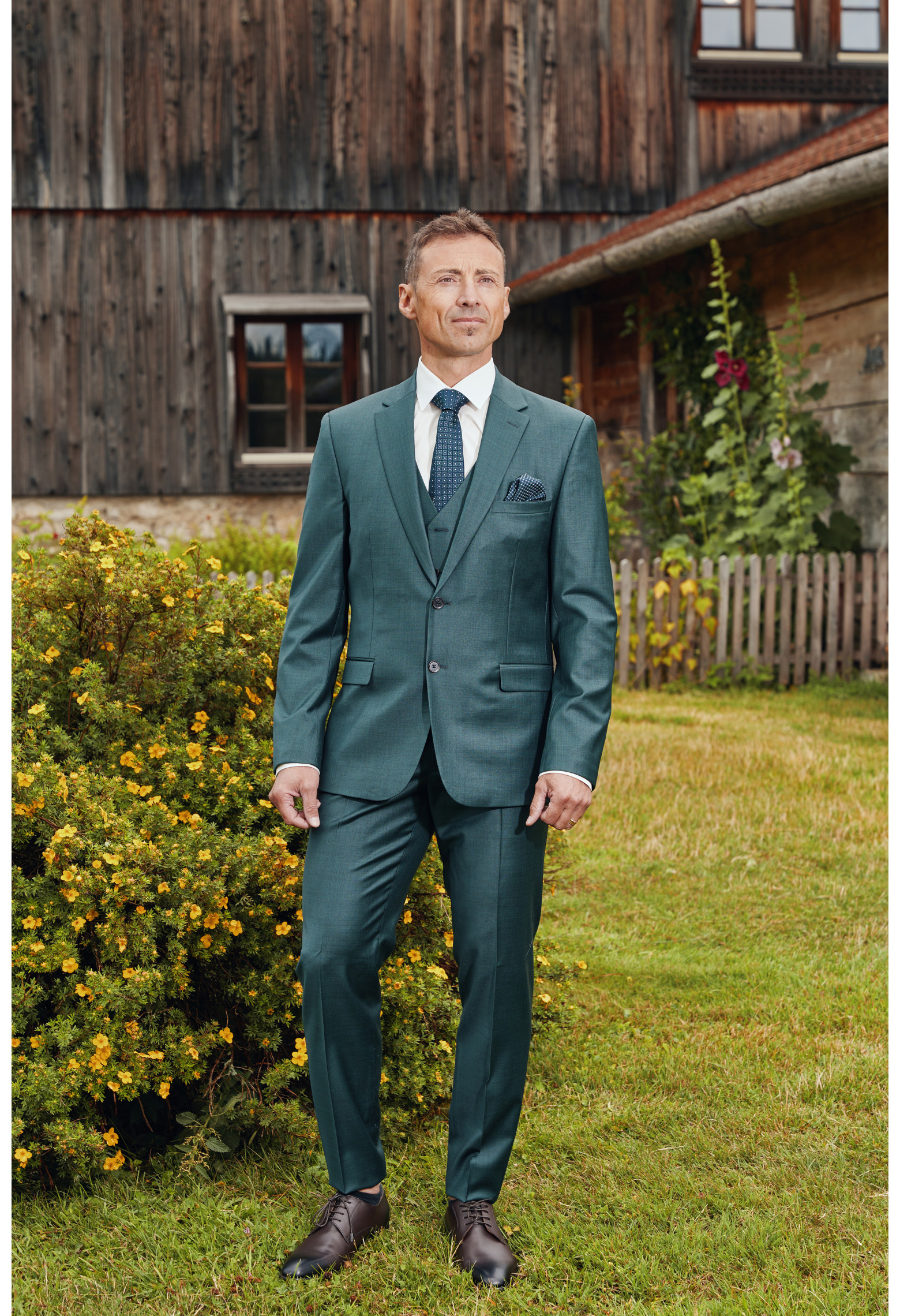 Homme de face en costume CITIZEN vert 2 pièces devant un chalet.