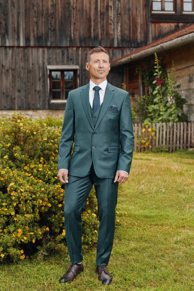 Homme de face en costume CITIZEN vert 2 pièces devant un chalet.