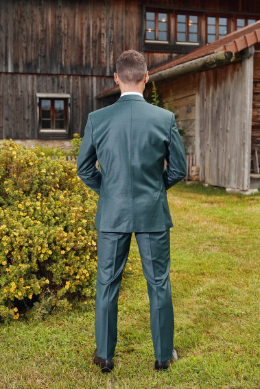 Homme de dos en costume CITIZEN vert devant un chalet . Il porte des chaussures marrons.