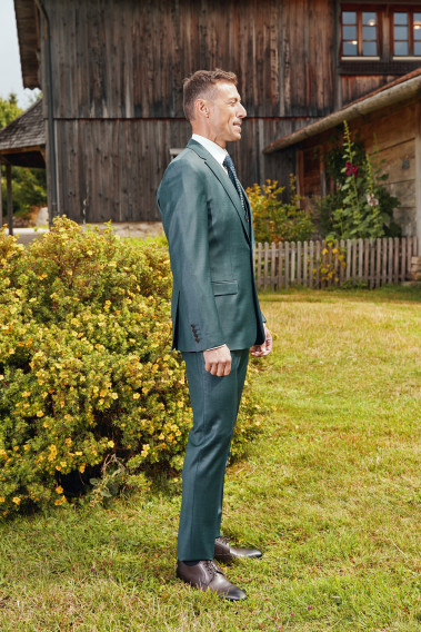 Homme de profil en costume CITIZEN vert devant un chalet . Il porte des chaussures marrons.