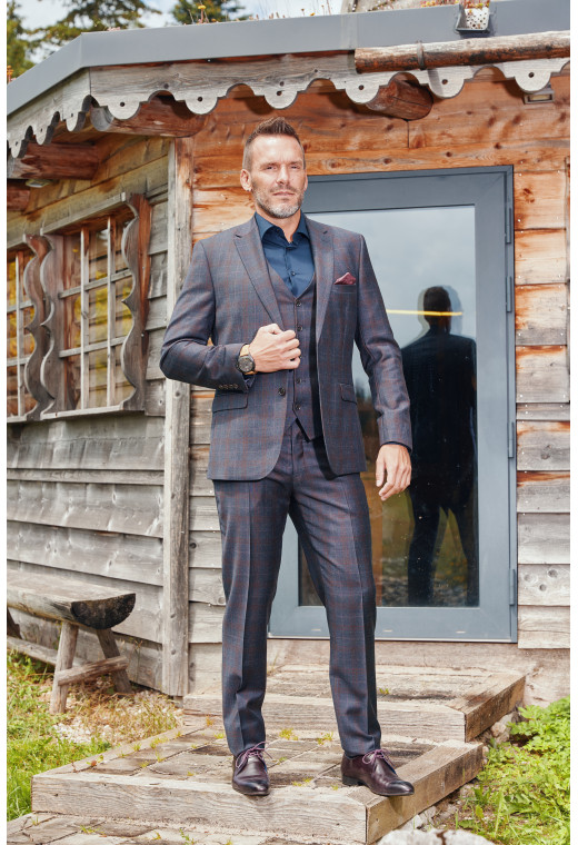 Homme de face en costume CITIZEN carreaux bordeaux 2 pièces devant un chalet.