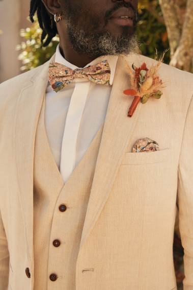 Costume GIORGIO de couleur beige. Zoom sur le nœuds  papillon et la poche à motif fleuri ainsi qu'un pins.