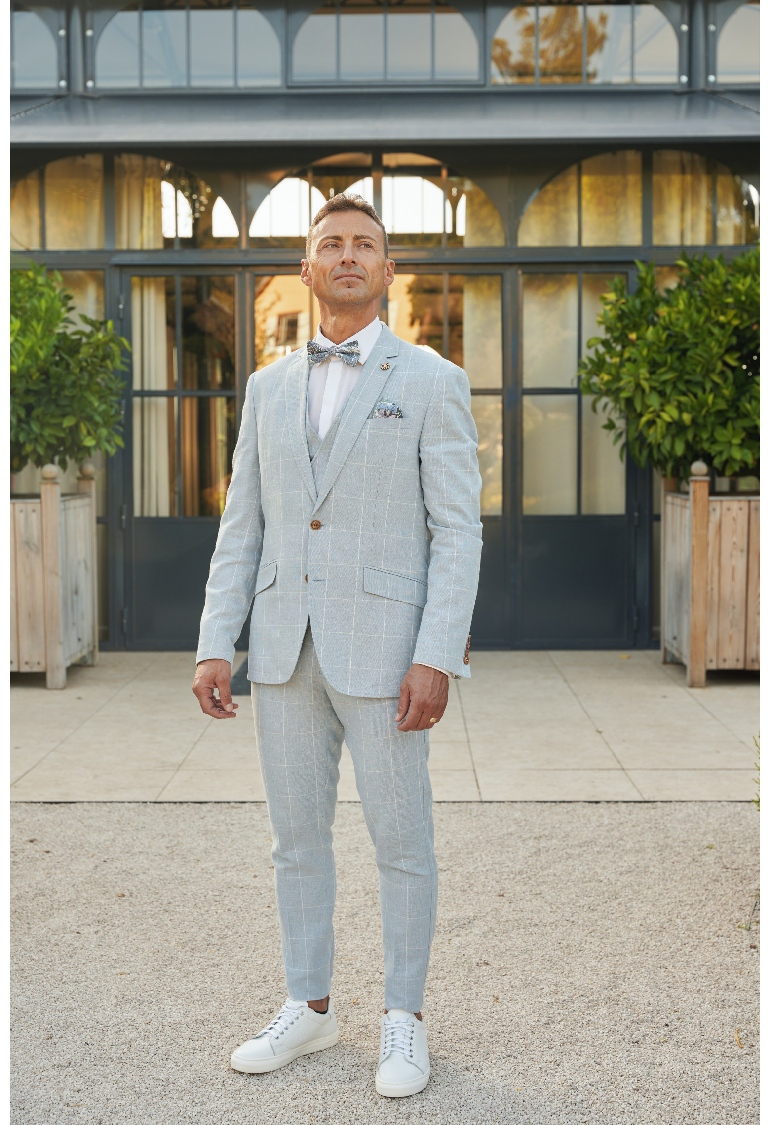 Homme de face en  costume GIORGIOC couleur bleu à carreaux 3 pièces devant une maison.