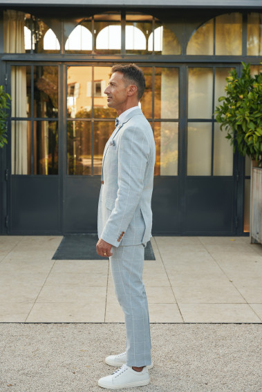 Homme de profil en  costume GIORGIOC couleur bleu à carreaux 3 pièces devant une maison.