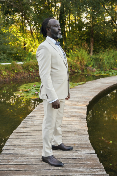Homme de profil en costume SUAT couleur craie 3 pièces sur un pont dans un lac. Il porte un nœuds papillon bleu foncé rayé.