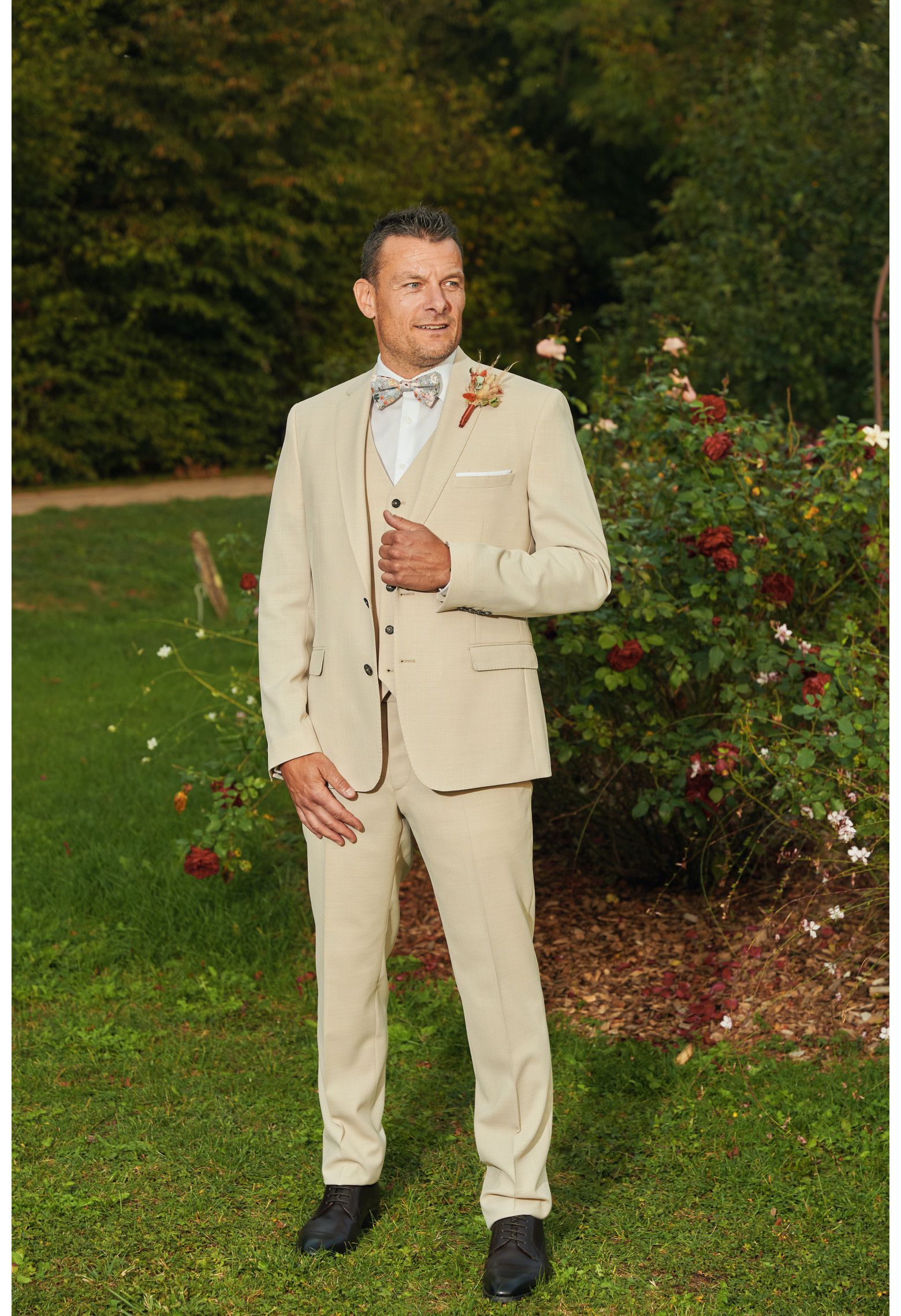 Homme de face en costume SUAT couleur beige 3 pièces dans une cours. Il porte un nœuds papillon et un pins fleuri.