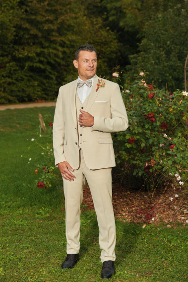 Homme de face en costume SUAT couleur beige 3 pièces dans une cours. Il porte un nœuds papillon et un pins fleuri.