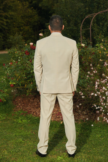 Homme de dos en costume SUAT couleur beige dans une cours. Il porte une chemise blanche et des chaussures marrons.