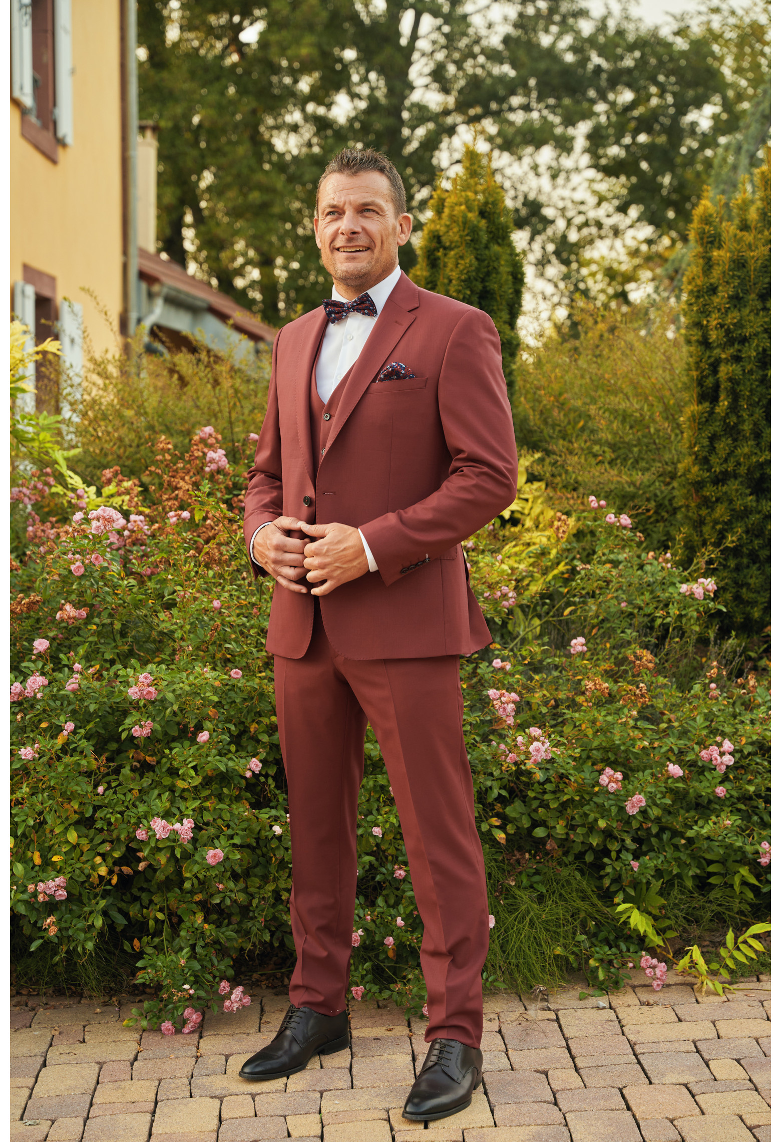 Homme de face en costume SUAT couleur framboise 3 pièces dans une cours fleurie.