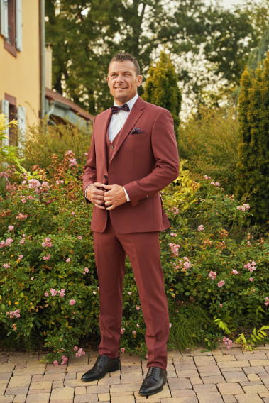 Homme de face en costume SUAT couleur framboise 3 pièces dans une cours fleurie.