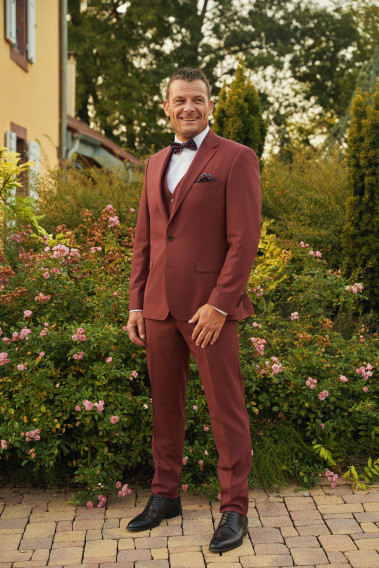 Homme de face en costume SUAT couleur framboise 3 pièces dans une cours fleurie.