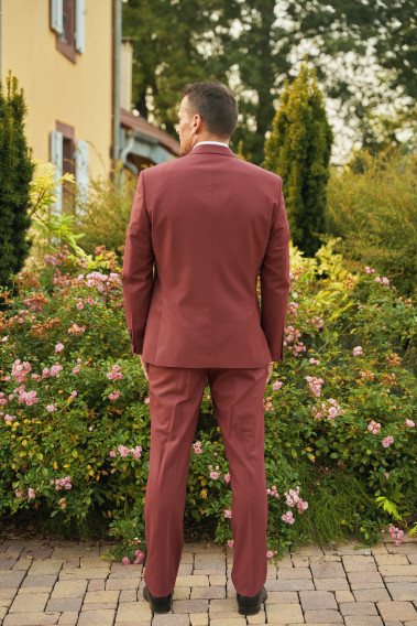 Homme de dos en costume SUAT couleur framboise 3 pièces dans une cours fleurie. Il porte des chaussures marrons.