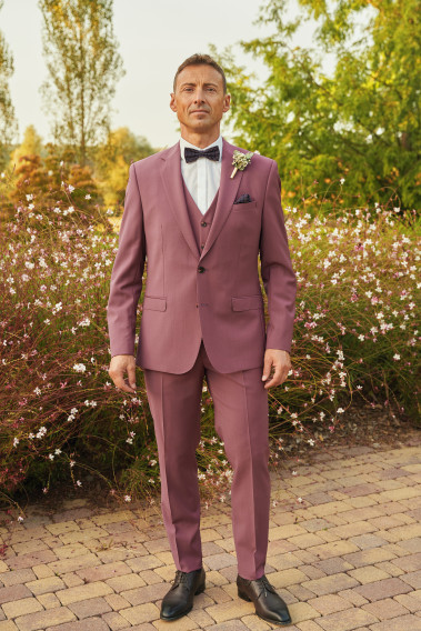 Homme de face en costume SUAT couleur fushia 3 pièces dans une cours fleurie.