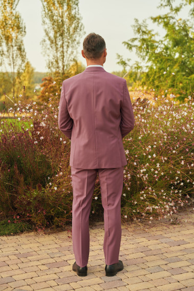Homme de dos en costume SUAT couleur fushia 3 pièces dans une cours fleurie. Il porte des chaussures marrons.