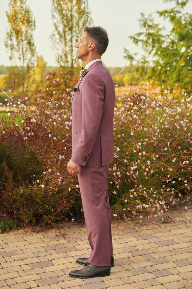 Homme de profil en costume SUAT couleur fushia 3 pièces dans une cours fleurie.