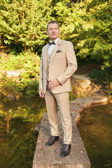 Homme en position trois-quarts en costume SUAT couleur beige sable 3 pièces sur un pont dans un lac.