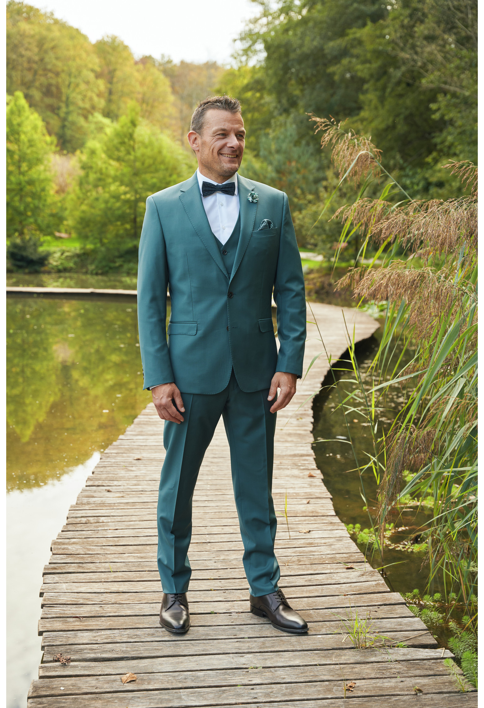 Homme de face en costume SUAT couleur vert bleuté 3 pièces sur un pont dans un lac.