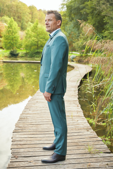 Homme de profil en costume SUAT couleur vert bleuté 3 pièces sur un pont dans un lac. Il porte des chaussures marrons.