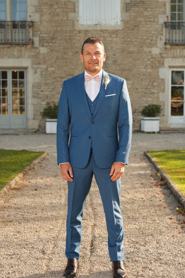 Homme de face en costume SUAT couleur bleu devant une maison.