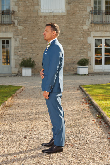 Homme de profil en costume SUAT couleur bleu 3 pièces devant une maison. Il porte des chaussures marrons.