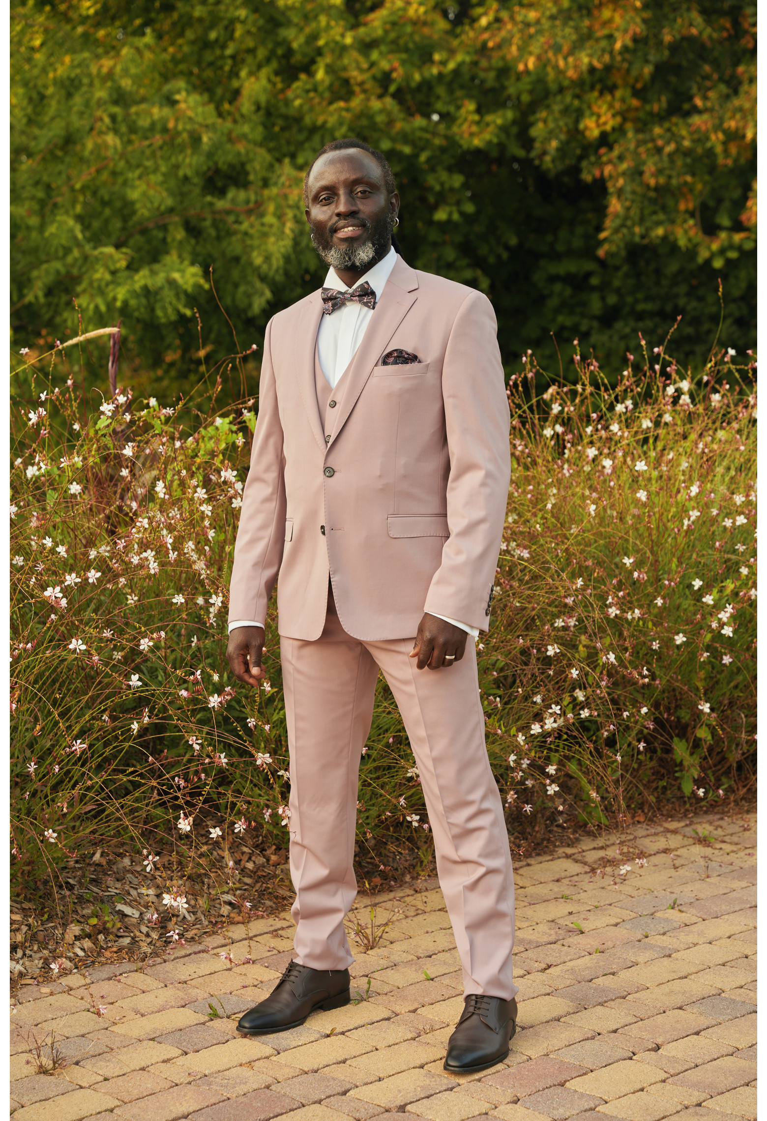Homme de profil en costume SUAT couleur rose pâle 3 pièces dans une cours fleurie.