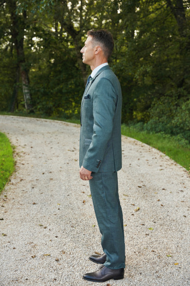 Homme de profil en costume SUAT couleur vert 3 pièces dans une cours. Il porte une cravate à motif.