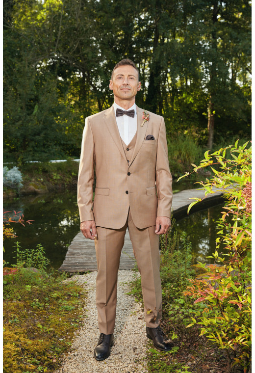 Homme de face en costume SUAT couleur beige foncé 3 pièces dans une cours avec un lac derrière.