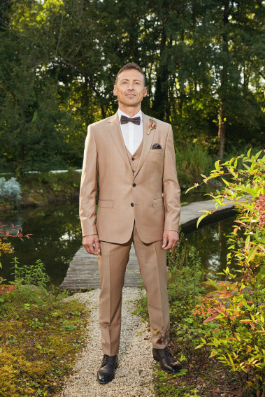 Homme de face en costume SUAT couleur beige foncé 3 pièces dans une cours avec un lac derrière.