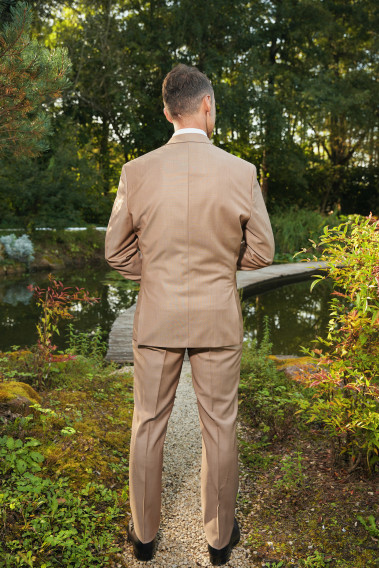 Homme de dos en costume SUAT couleur beige foncé dans une cours. Il porte une chemise blanche et des chaussures marrons.