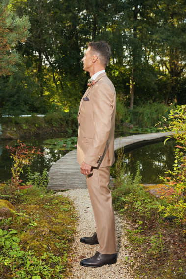 Homme de profil en costume SUAT couleur beige foncé 3 pièces dans une cours avec un lac derrière.